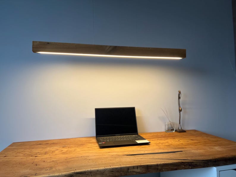 Może przedstawiać: Drewniana, prostokątna lampa wisząca z jasnym, liniowym źr&oacute;dłem światła LED. Światło wisi nad drewnianym biurkiem z laptopem i elementami dekoracyjnymi. Biurko wykonane jest z jasnego drewna.