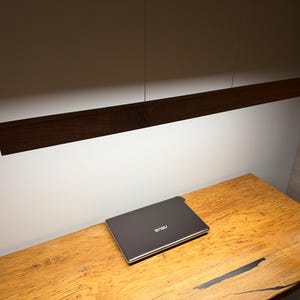 Może przedstawiać: Ciemnobrązowa drewniana liniowa lampa wisząca zawieszona nad drewnianym biurkiem. Zamknięty laptop znajduje się na biurku. Biurko ma wykończenie z naturalnego drewna.