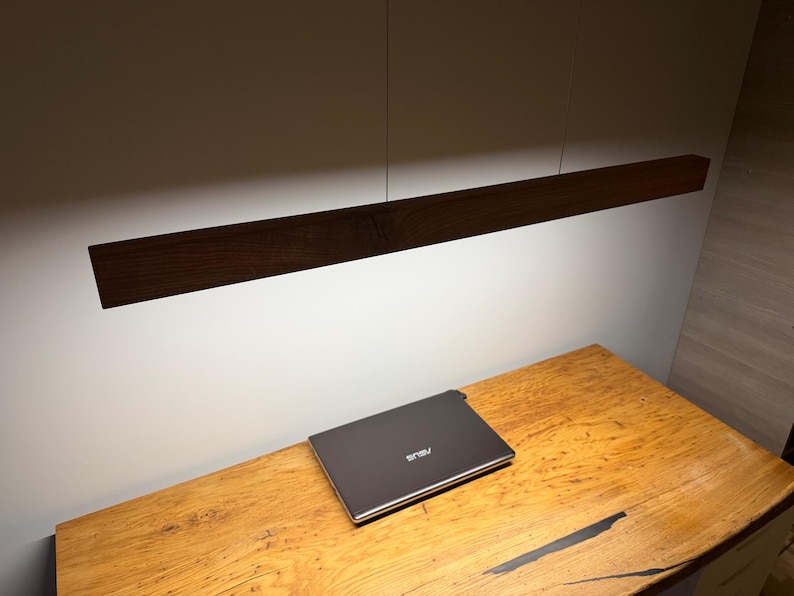 Może przedstawiać: Ciemnobrązowa drewniana liniowa lampa wisząca zawieszona nad drewnianym biurkiem. Zamknięty laptop znajduje się na biurku. Biurko ma wykończenie z naturalnego drewna.