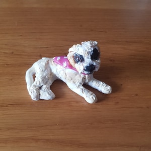 May include: A white and brown dog figurine with black eyes and a pink polka dot bandana. The figurine is lying down on a wooden surface.