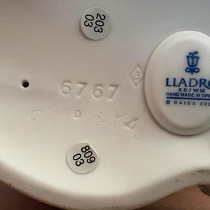 Pu&ograve; includere: La parte inferiore di una statuetta in porcellana bianca con il logo blu Lladro, il testo Hand Made In Spain, Daisa 2000 e X07WM. Il numero 6767 &egrave; in rilievo, insieme a due adesivi circolari che mostrano i numeri 203 03 e 809 03.