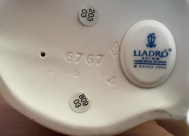 Pu&ograve; includere: La parte inferiore di una statuetta in porcellana bianca con il logo blu Lladro, il testo Hand Made In Spain, Daisa 2000 e X07WM. Il numero 6767 &egrave; in rilievo, insieme a due adesivi circolari che mostrano i numeri 203 03 e 809 03.