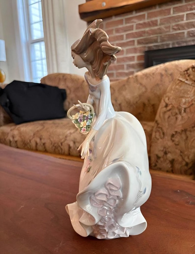 Pu&ograve; includere: Una statuetta in porcellana raffigurante una persona con capelli castani fluenti che indossa un lungo abito chiaro con dettagli a balze. La figura tiene un piccolo cestino pieno di fiori scolpiti colorati, con una finitura lucida.