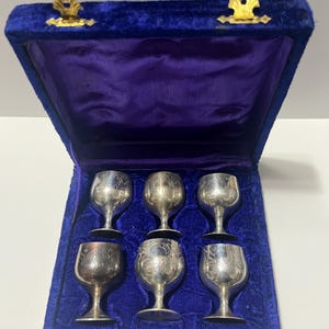 May include: A set of six silver goblets with etched floral patterns, displayed in a blue velvet-lined case with a purple interior. The case features gold-coloured clasps.
