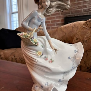 Pu&ograve; includere: Una statuetta in porcellana raffigurante una persona con lunghi capelli fluenti, che indossa un top azzurro e una lunga gonna bianca. La figura regge un cesto intrecciato pieno di piccoli fiori color pastello e presenta decorazioni floreali.