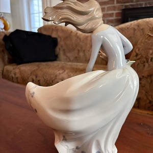 Pu&ograve; includere: Una statuetta in porcellana raffigurante una figura con lunghi capelli castano chiaro mossi dal vento, che indossa un top azzurro a maniche lunghe e un'ampia gonna bianca fluida. La gonna presenta delicati dettagli floreali blu sull'orlo, con una finitura liscia e lucida e un aspetto dinamico.