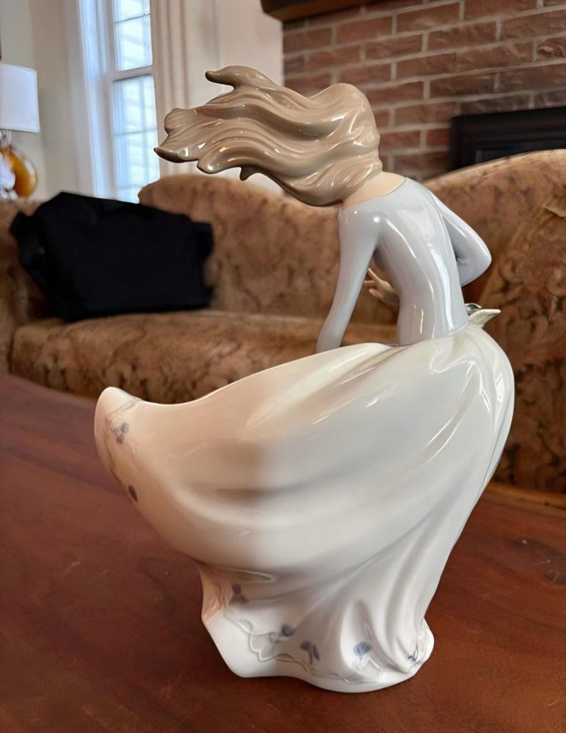 Pu&ograve; includere: Una statuetta in porcellana raffigurante una figura con lunghi capelli castano chiaro mossi dal vento, che indossa un top azzurro a maniche lunghe e un'ampia gonna bianca fluida. La gonna presenta delicati dettagli floreali blu sull'orlo, con una finitura liscia e lucida e un aspetto dinamico.