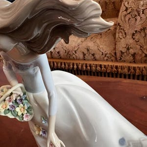 Pu&ograve; includere: Una statuetta in porcellana smaltata raffigurante una figura con lunghi capelli fluenti, che indossa un abito bianco. La figura tiene un cestino pieno di piccoli fiori multicolori e presenta ulteriori piccoli accenti floreali sulla gonna.