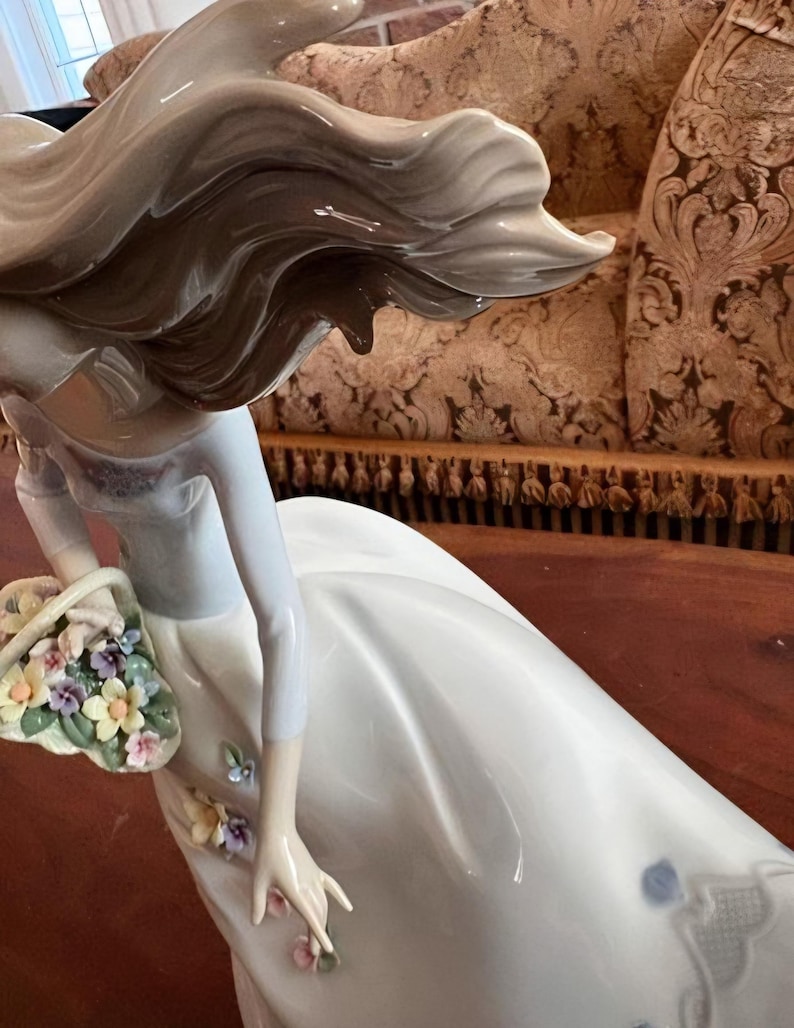 Pu&ograve; includere: Una statuetta in porcellana smaltata raffigurante una figura con lunghi capelli fluenti, che indossa un abito bianco. La figura tiene un cestino pieno di piccoli fiori multicolori e presenta ulteriori piccoli accenti floreali sulla gonna.