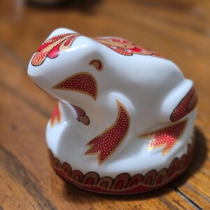 May include: A white ceramic frog figurine with red and gold decorative patterns. The frog has a rounded shape and is sitting on a wooden surface. The design includes a series of red dots and gold accents.