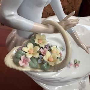 Pu&ograve; includere: Una statuetta in porcellana di una figura con un top azzurro che tiene in mano un cestino curvo pieno di fiori scolpiti. I fiori sono nelle tonalit&agrave; del giallo pallido, rosa tenue e azzurro, accentuati da foglie di colore verde.