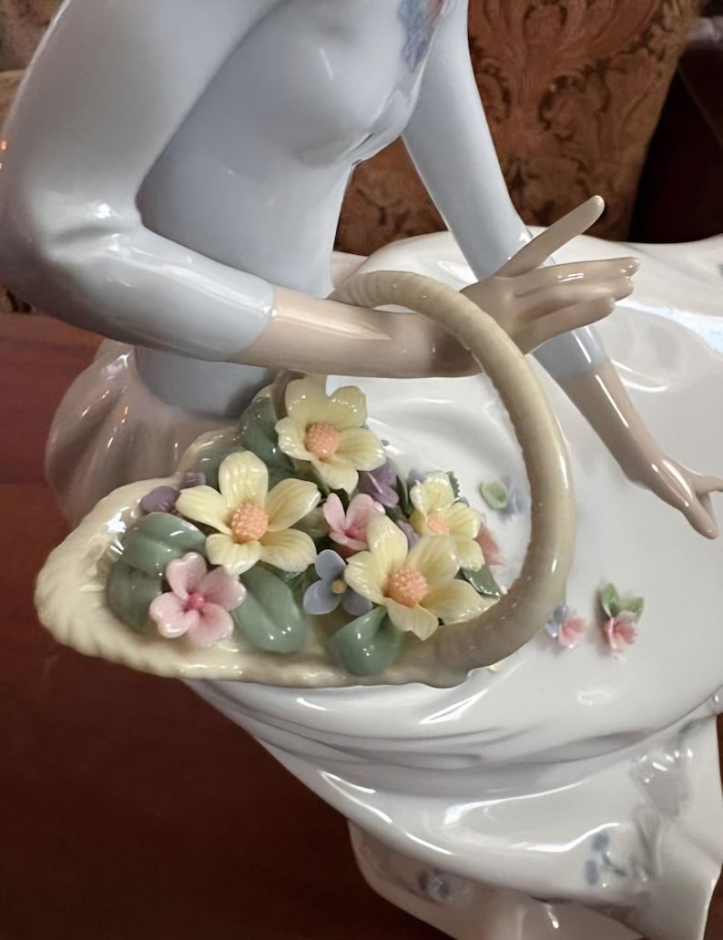 Pu&ograve; includere: Una statuetta in porcellana di una figura con un top azzurro che tiene in mano un cestino curvo pieno di fiori scolpiti. I fiori sono nelle tonalit&agrave; del giallo pallido, rosa tenue e azzurro, accentuati da foglie di colore verde.