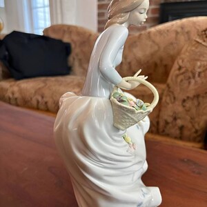 Pu&ograve; includere: Una statuetta in porcellana raffigurante una persona con capelli castano chiaro, che indossa un top a maniche lunghe azzurro e una lunga gonna bianca con dettagli floreali blu. La figura tiene in mano un cesto intrecciato pieno di fiori.