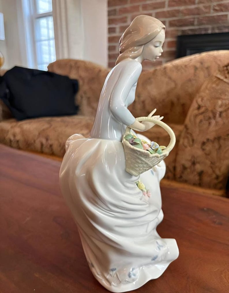 Pu&ograve; includere: Una statuetta in porcellana raffigurante una persona con capelli castano chiaro, che indossa un top a maniche lunghe azzurro e una lunga gonna bianca con dettagli floreali blu. La figura tiene in mano un cesto intrecciato pieno di fiori.