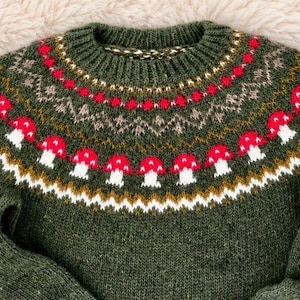 Könnte beinhalten: Ein dunkelolivgrüner Strickpullover mit Rundhalsausschnitt und einer Passe, die mit einem Fair-Isle-Muster verziert ist. Das Design zeigt Reihen von roten und weißen Pilzen, geometrischen Formen und Zickzacklinien in Weiß, Rot und Beige.