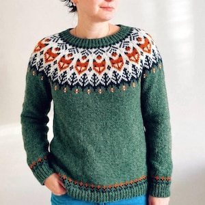 Woman wearing a Lyme Grass Green hand-knitted Icelandic wool Into the Wild lopapeysa sweater with a fox-pattern Nordic yoke in white, black and apricot . Traditional Icelandic lopapeysa featuring foxes and forest motifs, made from genuine Icelandic wool.