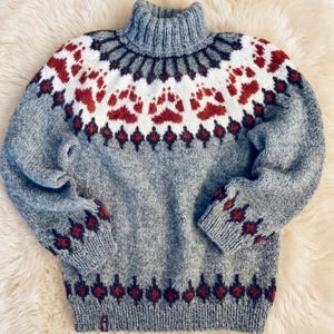 Può includere: Un maglione grigio lavorato a maglia con collo alto a lupetto. Il maglione presenta un motivo di impronte di zampe rosse su sfondo bianco, con forme a diamante rosse e blu scuro lungo il fondo e le maniche. Il maglione è realizzato con un filato morbido e strutturato.