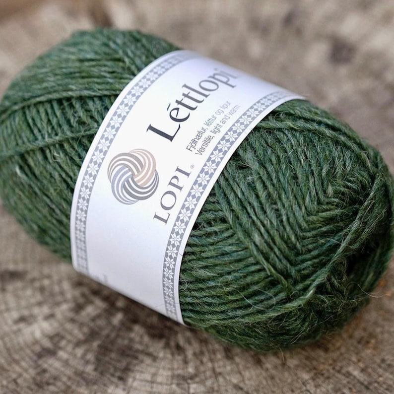 May include: A ball of dark forest green L&egrave;ttlopi yarn wrapped in a white paper label. The label features the text L&egrave;ttlopi and Lopi, a circular logo, and the phrase Versatile, light and warm, framed by a decorative border of small white snowflakes.
