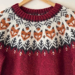 Close-up of a red hand-knitted Into the Wild Icelandic wool lopapeysa sweater with an Apricot fox-pattern yoke in white and dark gray. Nordic lopapeysa featuring foxes and forest motifs, hand-knitted from genuine Icelandic wool.