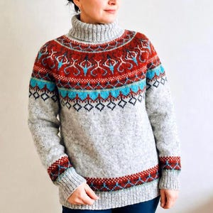 Può includere: Un maglione in maglia grigio chiaro con collo alto a coste e maniche lunghe. Il carré e l'orlo sono decorati con un colorato motivo fair isle nelle tonalità del rosso intenso, turchese brillante e nero, creando un design geometrico.