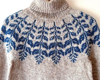 Può includere: Un maglione a collo alto beige con un motivo geometrico blu sulla parte superiore. Il maglione è realizzato in materiale a maglia e viene esposto su una gruccia di legno. Il design presenta forme a diamante e a foglia.
