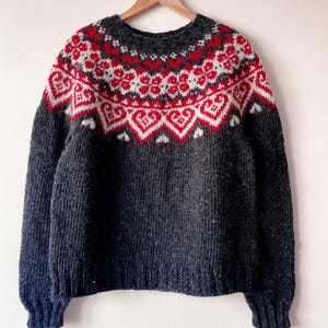 May include: A charcoal gray knit sweater featuring a round neckline and long sleeves. The yoke displays a detailed pattern of red and white hearts and floral motifs. The sweater is constructed with a ribbed collar, cuffs, and hemline.