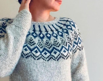 May include: A light gray long-sleeve sweater featuring a round neckline and a dark blue and white geometric pattern across the yoke and chest. The knit garment has a textured appearance with a ribbed collar and cuffs, showcasing a classic fair isle style design.
