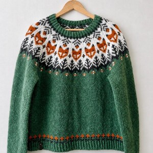 May include: A green long-sleeve sweater featuring a circular yoke with a repeating pattern of orange fox faces, white geometric shapes, and black pine tree motifs. The hem and cuffs are ribbed, and a small orange and black border accents the bottom.