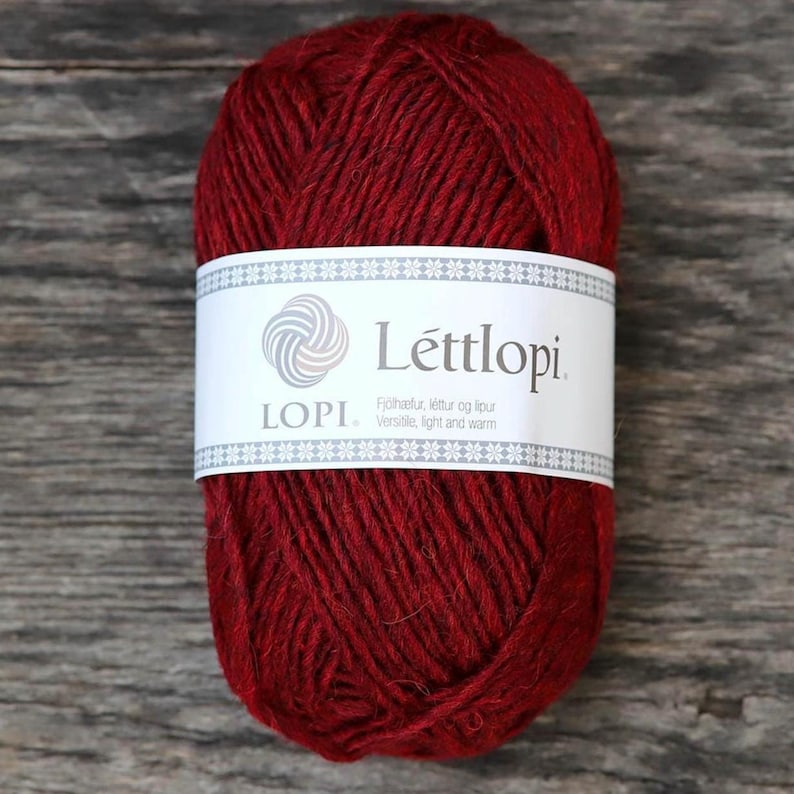 May include: A skein of deep red yarn with a white paper label wrapped around the centre. The label features the text L&eacute;ttlopi, LOPI, and the phrase Versatile, light and warm, along with a circular logo and a decorative border of small stars.