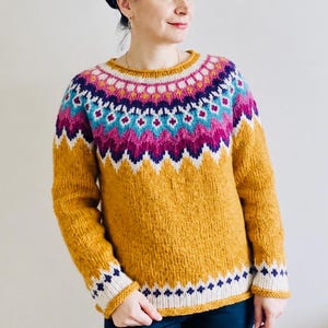 May include: A long-sleeved knit sweater featuring a mustard yellow base with a vibrant yoke pattern. The yoke displays horizontal bands of white, magenta, dark blue, and turquoise in a geometric design. The cuffs and hem have white trim.