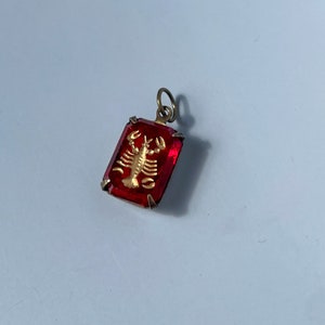 May include: A gold-colored zodiac charm pendant featuring the Cancer sign, a crab, set in a red glass stone.