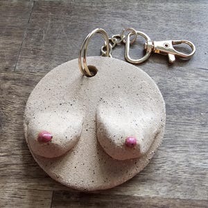 Puede incluir: Un llavero de cerámica circular de color beige moteado que presenta dos relieves en forma de pecho con pequeños pezones pintados en rosa brillante. La pieza de cerámica está unida a una anilla de metal dorado y un cierre de mosquetón.