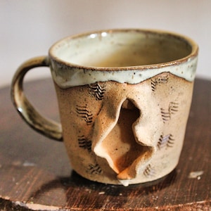 May include: A ceramic mug with a speckled tan finish and a smooth, light-colored glaze along the top rim. The exterior features a central, vertically oriented, folded clay protrusion and small, stamped chevron patterns across the surface.