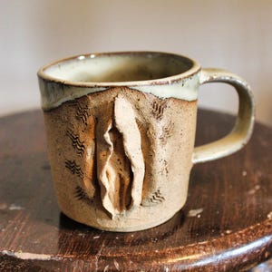 May include: A handmade ceramic mug with a speckled tan finish and a smooth, light-colored glazed rim. The exterior features a central, raised, vertical organic sculptural element surrounded by small, stamped, wavy line patterns on both sides.