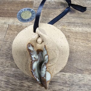 Peut inclure: Un ornement en céramique beige moucheté de forme circulaire, présentant une sculpture anatomique centrale avec un intérieur émaillé brun et crème brillant. Un ruban noir est passé dans un trou au sommet pour la suspension. Une pièce de deux dollars canadiens est visible à proximité.