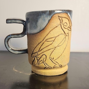 Op de afbeelding: Een handgemaakte keramische mok met een uniek vogelontwerp. De mok heeft een bruin lichaam met een uitgesneden vogelillustratie en een donkerblauwe rand en handvat. De mok is ongeveer 10 cm hoog.