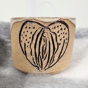Peut inclure: Une tasse en céramique faite à la main avec un motif en forme de cœur. La tasse est de couleur marron clair avec un motif de cœur marron plus foncé sculpté sur le devant. Le bord de la tasse est de couleur beige clair.