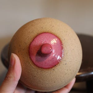 May include: A spherical ceramic object with a speckled tan, matte finish. The centre features a circular, glossy, dark pink glazed area with a small, raised, cylindrical protrusion in the middle, creating a textured and contrasting appearance.