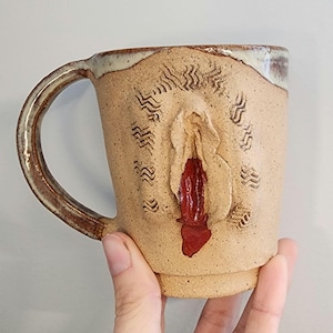 Puede incluir: Una taza de cerámica hecha a mano con exterior beige y asa marrón. La taza presenta un diseño de vulva esculpida con detalles en rojo. La taza está sostenida por una mano.