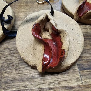 Peut inclure: Un ornement circulaire en céramique beige avec une texture mouchetée et un élément central en argile pliée et surélevée, émaillé d'un rouge profond brillant. Un ruban noir passe dans le disque et un fermoir mousqueton doré est fixé.