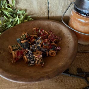 May include: A collection of small wooden spools wrapped in various plaid and checkered fabric strips, resting inside a large, round, dark brown wooden bowl. The fabric patterns feature red, blue, green, and white colors in different combinations.
