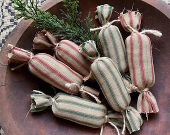 May include: A wooden bowl filled with fabric candy decorations. The cylindrical ornaments are tied with twine and display red and green stripes on a beige fabric. A sprig of greenery is placed amongst the ornaments.