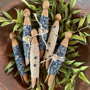 May include: A group of six wooden clothespins, each wrapped in a different blue and white patterned fabric. The clothespins are arranged in a wooden bowl with green leafy foliage.