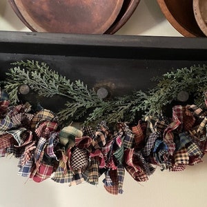 May include: A plaid fabric garland with a green garland of pine branches draped over a black shelf.