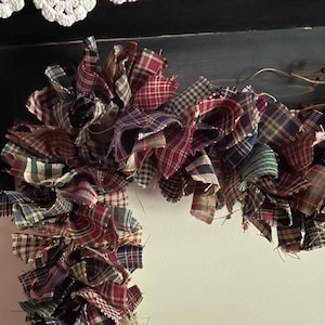 May include: A decorative candy cane shaped ornament constructed from numerous strips of fabric featuring various plaid patterns in shades of red, blue, green, and tan. The fabric pieces are tied together to create a full, textured, ruffled appearance.