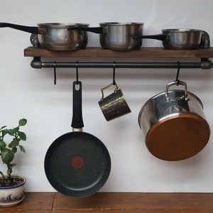 May include: A rustic wooden shelf with black metal pipe supports and hooks. The shelf holds four stainless steel pots and a black frying pan. A gold mug hangs from one of the hooks.