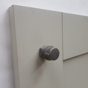 May include: A close-up of a grey metal plug screwed into a light grey wooden panel. The plug has a hexagonal shape and the number "1/2" is visible on its top. The panel is made of two connected pieces.