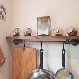 May include: A wooden shelf with a metal pipe rail and hooks. There are five glass jars on the shelf, four of which contain spices. The fifth jar is empty. There are three black frying pans hanging from the hooks.
