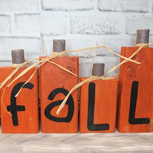May include: Wooden pumpkin-shaped blocks spell out the word "FALL" in black paint. The blocks are painted orange and have a rustic finish. Each block is tied with natural twine.
