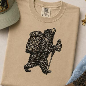 May include: A beige crew neck t-shirt featuring a black graphic print of a bear wearing a large backpack and holding a hiking pole and a water bottle. The shirt has a Comfort Colors brand label at the neck indicating a size large, with a crew neckline.